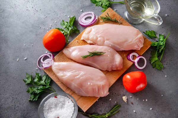 pieces of raw chicken fillet on a cutting Board, meat, vegetables, herbs, tomato, onion, parsley, oil, salt on a gray background