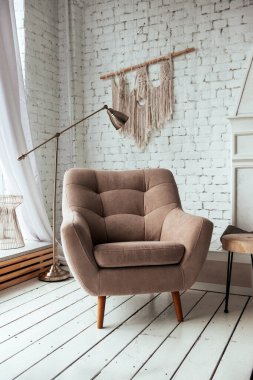 İçi aydınlık bir oda, beyaz ahşap bir zeminde bej bir koltuk, pencere, lamba