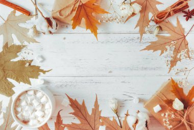 Sıcak çikolata, 1 fincan marşmelovlu kakao, sonbahar sarısı akçaağaç yaprakları, koniler, tarçın çubukları, bir hediye kutusu, beyaz çiçekli bir dal ve beyaz ahşap arka planda pamuk.