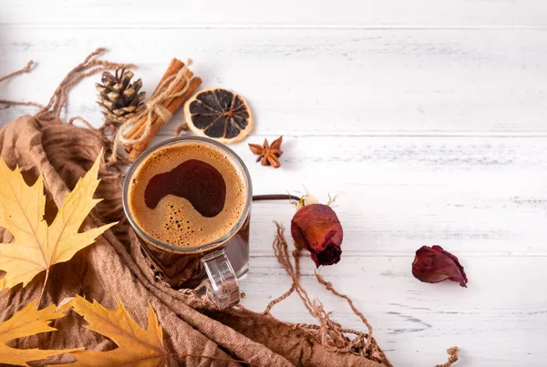 Bir fincan sade sıcak kahve, sonbahar sarısı akçaağaç yaprakları, koniler, tarçın çubukları, kahverengi bir eşarbın yanında kurumuş gül, beyaz ahşap bir arka planda bir battaniye.