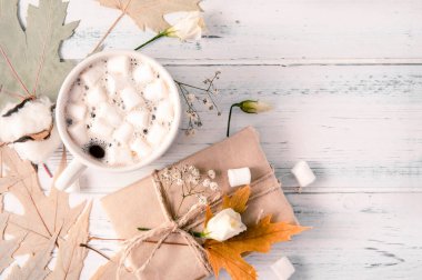 1 kupa kremalı sıcak çikolata, akçaağaç sarısı kuru yapraklar, hediye kutusu, beyaz tahta üzerinde beyaz çiçekler. 