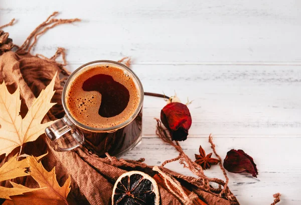 Kahverengi eşarplı, akçaağaç yapraklı, tarçınlı, kurumuş güllü, beyaz ahşap arka planda bir dilim kuru portakallı bir fincan sade kahve.