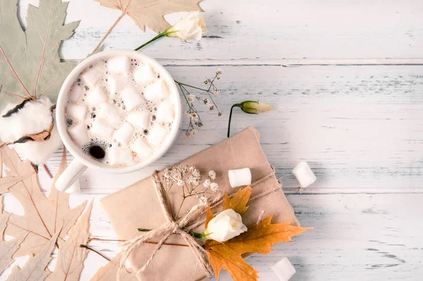 1 kupa kremalı sıcak çikolata, akçaağaç sarısı kuru yapraklar, hediye kutusu, beyaz tahta üzerinde beyaz çiçekler. 