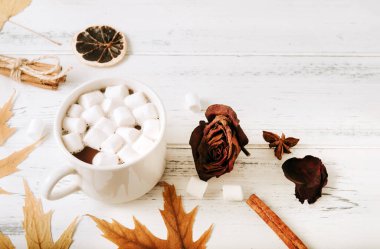 1 fincan kremalı sıcak çikolata, akçaağaç kuru yaprakları, tarçın çubukları, kurutulmuş gül, beyaz ahşap arka planda bir dilim kuru portakal.