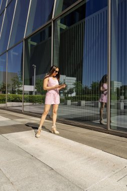 Modern ofis binasının önünde akıllı telefon kullanan iş kadını cam cepheye yansıyor.