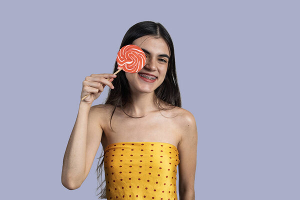 Студийный портрет веселой молодой женщины, закрывающей один глаз леденцом, носящей зубные брекеты и улыбающейся
