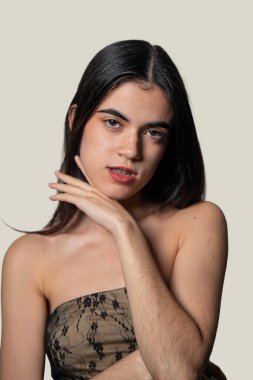 Uzun siyah saçlı ve diş telli genç bir Latin kadının stüdyo portresi, elleri çenesinde poz verirken vücut kıllarını güvenle sergiliyor.