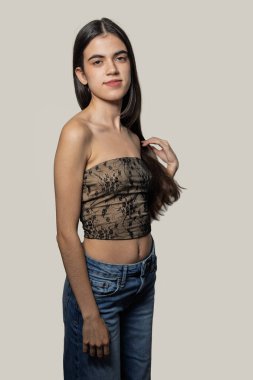 Saçına dokunan, askısız bluz ve kot giyen genç bir kadının stüdyo portresi.