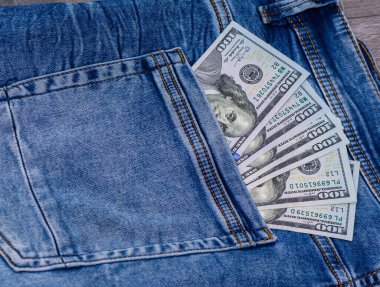 100 ABD banknotu bir kot pantolon cebinde kısmen görünür, parayı, mali güvenliği, tasarrufu ve günlük harcamaları temsil eder.