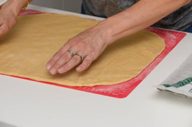 Bir kadın hamuru un bir masanın üzerinde, ahşap bir oklamalı iğne ile yuvarlıyor, sıcak bir ev mutfağında ev yapımı hamur işleri hazırlıyor.