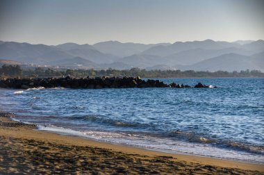 Kuzey Kıbrıs 'ta bir plajın üzerinde gün batımı manzarası ve uzak dağlar. Akşam gökyüzünün ılık renkleri sakin deniz suyuna yansıyor.