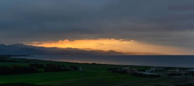 Güneş ufkun altına batarken, altın ışınlar engebeli kıyı dağlarını ve sakin denizi aydınlatıyor. Üzerimizde kara bulutlar sürükleniyor, akşam gökyüzünde dramatik bir kontrastlık yaratıyor..