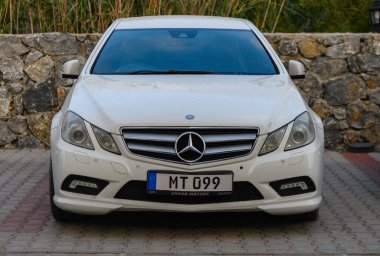 Otoparka park etmiş beyaz bir Mercedes-Benz E-Sınıfı sedan..