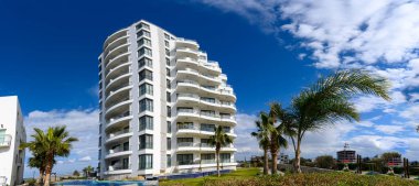 Çarpıcı bir modern bina, kabarık bulutlarla süslenmiş canlı mavi bir gökyüzüne karşı dimdik ayakta duruyor. Görkemli palmiye ağaçları sahneyi çerçeveleyerek, konumun tropikal atmosferini güçlendirir.