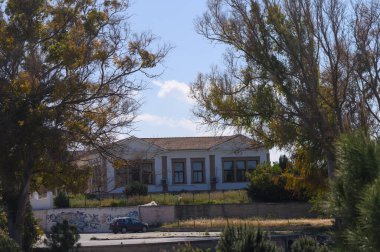 Eski, terk edilmiş bir bina uzun ağaçların arasında sessiz duruyor, solgun boya tarihini açığa vuruyor. Yukarıdaki mavi gökyüzü yalnızlık duygusunu artırıyor. Zamanla unutulan bir şehir ortamını yakalıyor..