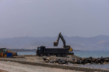 Kazıcı, Kıbrıs Rum Kesimi 'ndeki Akdeniz kıyısı inşaatı sırasında büyük deniz taşlarını bir çöp kamyonuna taşıyor.
