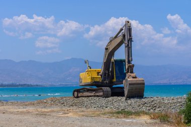 Kıbrıs 'ta güneşli bir plajda çalışan sarı bir kazıcı, turkuaz Akdeniz yakınlarındaki kumu düzleştiriyor. Parlak yaz günü, inşaat sahnesi.