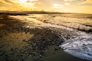 Gün batımında sakin bir sahil, ılık renklerin gökyüzünü kucakladığı ve dalgaların çakıl taşlı kıyıya hafifçe vurduğu..
