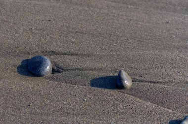 Deniz taşları ve çakıl taşları, sakin bir yaz manzarası yaratan yumuşak Akdeniz dalgaları ve güneşli gökyüzü ile bir Kıbrıs plajının kumlu sahillerine saçılmış..