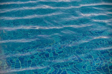 Yüzme havuzundaki berrak mavi su yüzeyinde güneş ışığı yansımaları var..