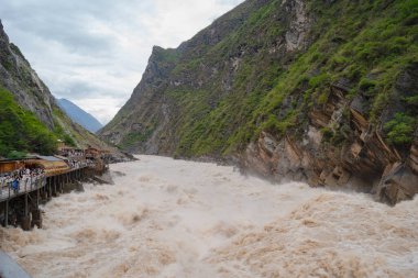 Turistler, insanların derin, yeşil bir geçitten geçen azgın, çamurlu nehir manzarasının keyfini çıkarıyorlar. Jinsha Nehri. Çin 'in en derin kanyonları.. 