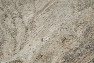 Yalnız bir yürüyüşçü, çorak ve kayalık bir dağ manzarasında bir patikada yürüyor..