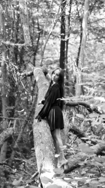 Siyah elbiseli genç bir kadın ormanda çıplak ayakla dikiliyor, yeşil yapraklar ve doğal güneş ışığıyla çevrili bir ağaca yaslanıyor. Doğayla uyum, özgürlük ve doğal güzellik kavramı..