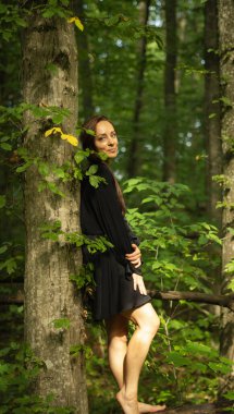 Siyah elbiseli genç bir kadın ormanda çıplak ayakla dikiliyor, yeşil yapraklar ve doğal güneş ışığıyla çevrili bir ağaca yaslanıyor. Doğayla uyum, özgürlük ve doğal güzellik kavramı..