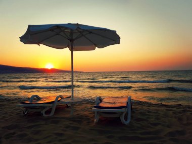 İki şezlong ve şemsiye güneş ışınları önünde Kum Plajı ile deniz sahilinde bir gün batımı olarak romantik bir sahne.