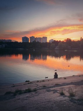 Huysuz ve huzurlu bir gece sahnesi. Küçük bir kız silueti, vahşi ördekleri besliyor, parktaki gölün kenarında oturuyor. Sakin su yüzeyinde rengarenk bir yansımayla harika bir yaz günbatımı..
