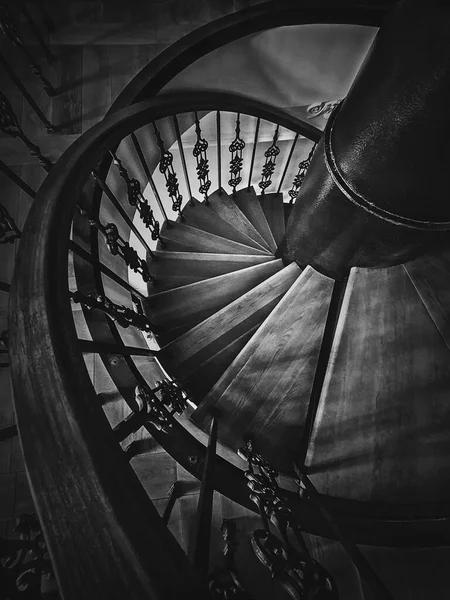 A look down to an old spiral staircase. Wooden circular stairway with ornate metallic railing, black and white vertical shot, interior architecture details of stair steps inside an ancient building.