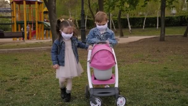 Kleine lustige Kinder in medizinischen Masken spielen auf dem Spielplatz mit dem Kinderwagen.