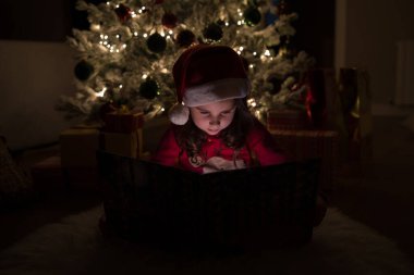 Noel pijaması giyen ve Noel Baba şapkalı bir kız Noel ağacının altında hediye dolu bir kitap okuyor.