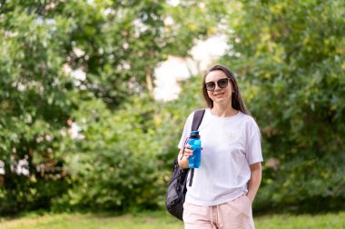 Aktif bir kadın, sıcak ve güneşli bir günde parkta sırt çantası ve spor şişesiyle susuzluk ve temiz havanın tadını çıkarıyor.