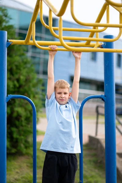 Odaklanmış sarışın çocuk açık hava antrenmanı sırasında yatay barlara takılır, kuvvet kazanır ve şehir parkında eğlenir.