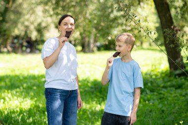 Mutlu anne ve oğul, güneşli yaz parkında birlikte çikolatalı dondurmanın tadını çıkarıyorlar. Aile eğlencesi ve açık havada tatlı ikram kavramı.