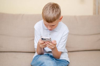 Genç çocuk bir kanepede oturuyor, akıllı telefonuna dalmış, modern alet bağımlılığını ve gençler arasındaki aşırı ekran zamanını resmediyor.