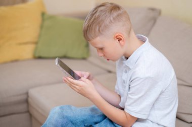 Genç çocuk bir kanepede oturuyor, akıllı telefonuna dalmış, modern alet bağımlılığını ve gençler arasındaki aşırı ekran zamanını resmediyor.