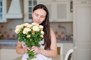 Uzun saçlı genç bir kadın mutfakta Anneler Günü 'nde hediye edilen sarı gül buketinin keyfini çıkarıyor.