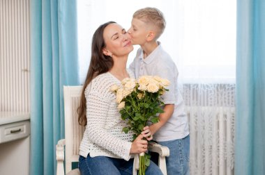 Ergen çocuk sıcak bir odada anneler gününü sevgi ve minnettarlıkla kutlayan sarı gülleri sunarken annesine sıcak bir şekilde sarılıyor.