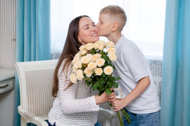 Ergen çocuk sıcak bir odada anneler gününü sevgi ve minnettarlıkla kutlayan sarı gülleri sunarken annesine sıcak bir şekilde sarılıyor.