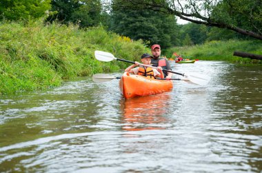 Baba ve oğul güneşli bir yaz gününde sakin, manzaralı bir nehirde kanoya biniyor, açık hava macerasının, doğa, su sporunun, aile aktivitesinin keyfini çıkarıyorlar.