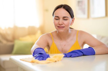 Eldivenli neşeli bir kadın masayı bezle siliyor, temizliği kucaklıyor ve bir gülümsemeyle evde bakım yapıyor.