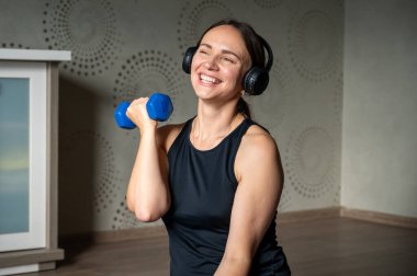 Kulaklık takan bir kadın evde dambıllarıyla egzersiz yapıyor, güç antrenmanı yapıyor ve online spor yapıyor, sağlıklı yaşam tarzı kavramından keyif alıyor.