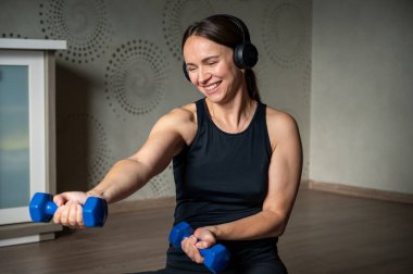 Kulaklık takan bir kadın evde dambıllarıyla egzersiz yapıyor, güç antrenmanı yapıyor ve online spor yapıyor, sağlıklı yaşam tarzı kavramından keyif alıyor.