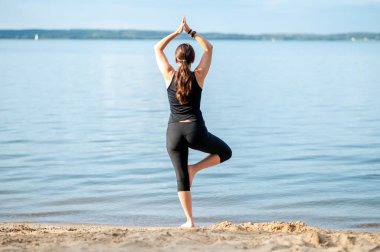 Siyah yoga kıyafetleri içindeki güzel genç kadın yazın büyük bir gölün kıyısında poz veriyor, doğanın tadını çıkarıyor ve sağlıklı yaşam tarzını geliştiriyor.