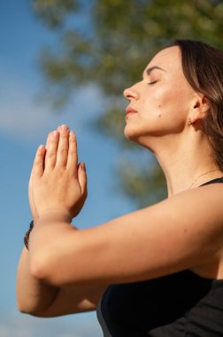 Dalgalı saçlı genç bir kadın açık havada meditasyon yapıyor güneşli bir günde, elleri dua pozunda katlanmış, dingin ve canlı bir doğal çevrede sakin ve bilinçli bir şekilde.
