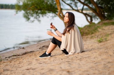 Battaniyeye sarılı genç bir kadın göl kıyısında oturur, çayını yudumlar ve çikolata yer, rahatlatır ve doğada rahat bir anın tadını çıkarır.