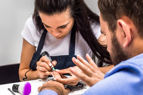 Young woman doing manicure to a man. A man in a shirt and with a beard looks at his hand.