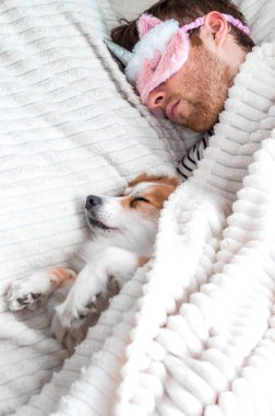 Closeup portrait of a young man in a pink mask sleeping in a bed under a rug with his dog. Vertical photo